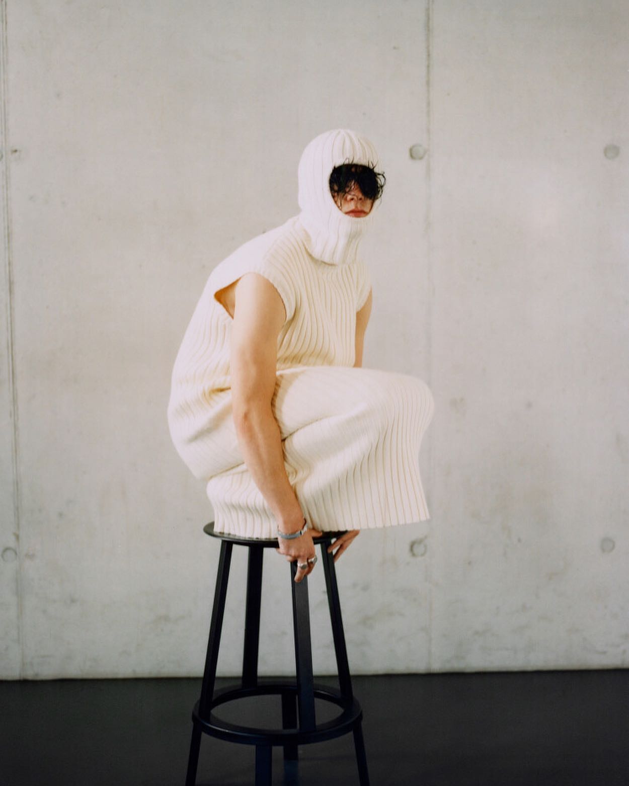 An adult wearing a cream-colored, knitted balaclava and matching clothing squats on a black stool against a pale wall.