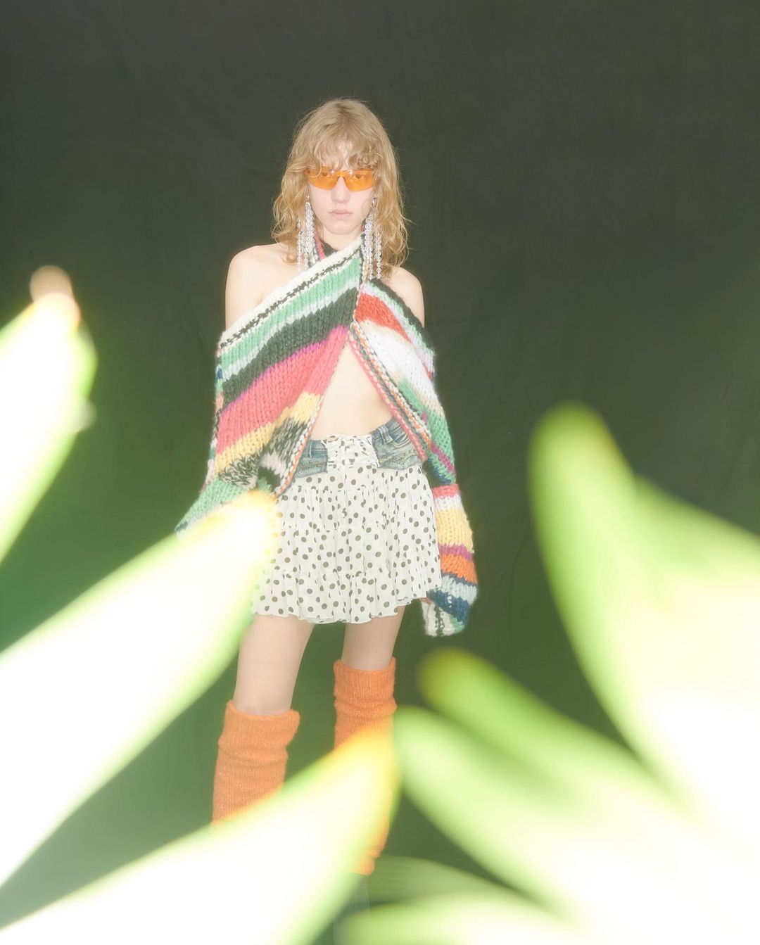 A woman with long hair in sunglasses poses in a colorful sweater, polka dot skirt, and leg warmers with greenery in the foreground.