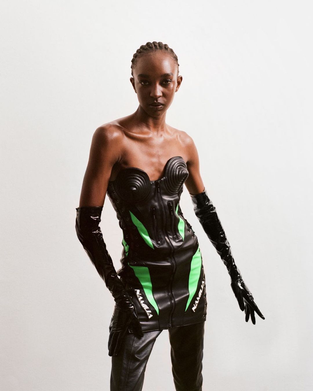 Fashion shot of a slender Black female model in a black futuristic bodysuit with neon green accents, black gloves, and black pants posing.