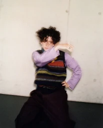 Person with dark curly hair wearing a striped knit vest over a lavender shirt against a concrete wall