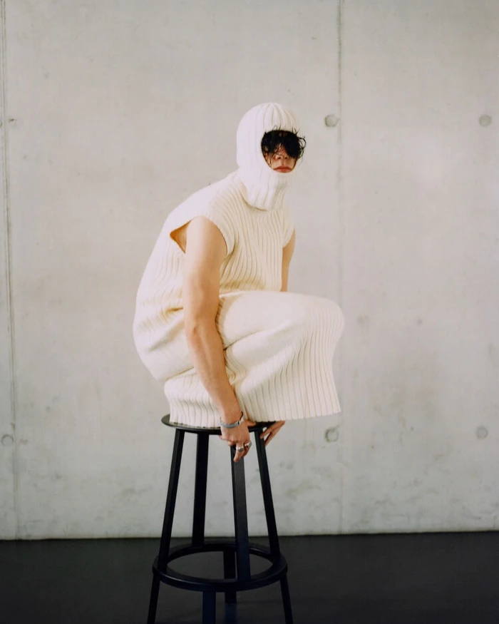 Model in cream ribbed knit balaclava and sleeveless tunic crouched on a black stool against concrete