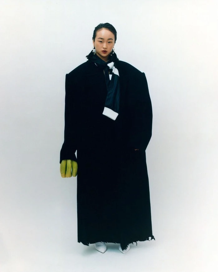 Model in oversized black coat holding green bananas against a plain white background