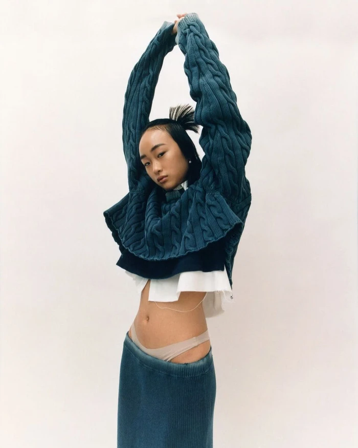 Model in blue cable knit sweater and denim skirt with arms raised against a plain white studio background