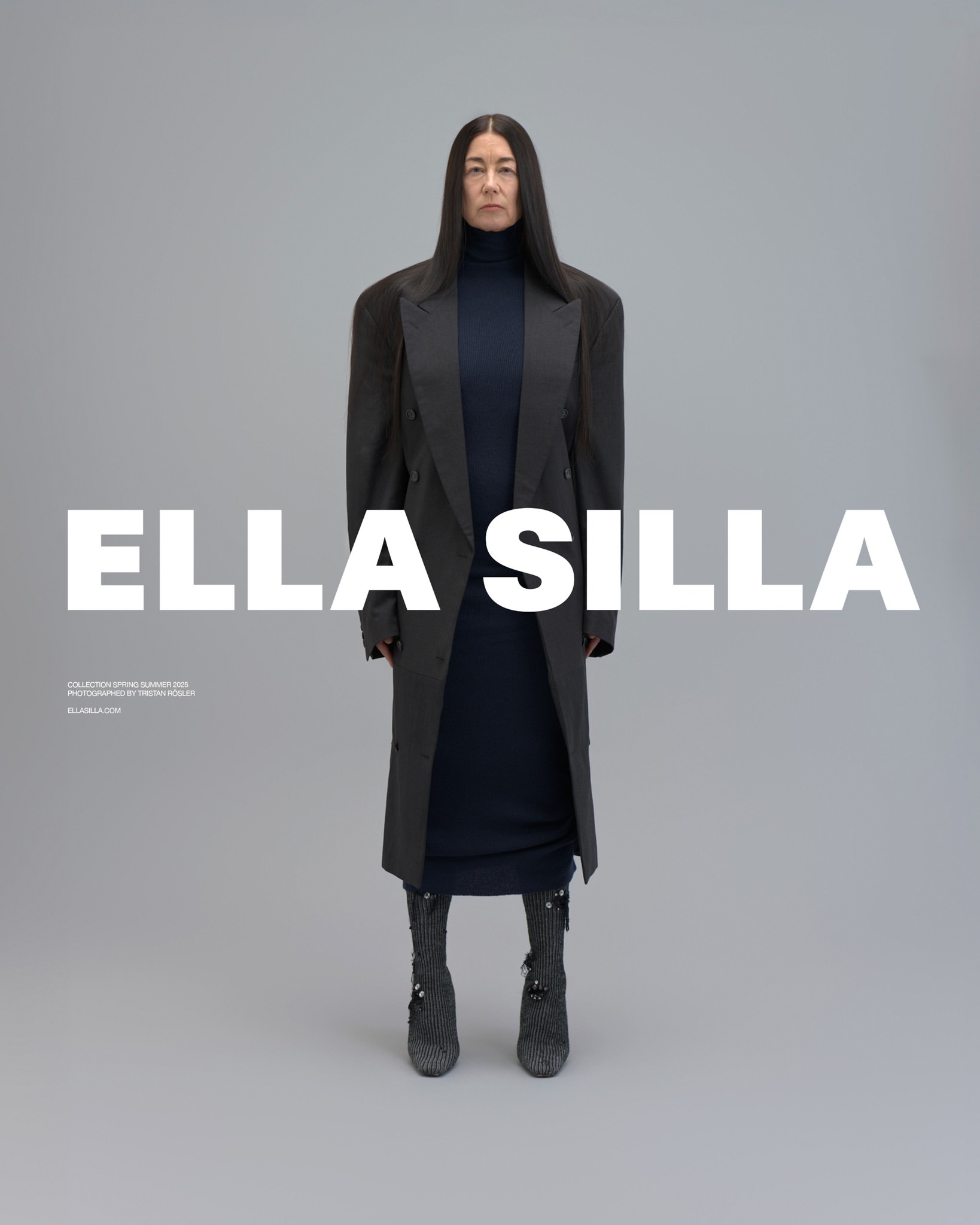 Model with long dark hair wearing a dark oversized coat and turtleneck dress standing against a grey studio backdrop