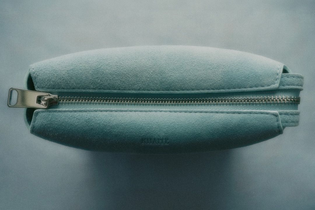 Close-up shot of a light blue suede cosmetic bag with silver zipper hardware, showcasing the fabric's texture and the design's clean lines.