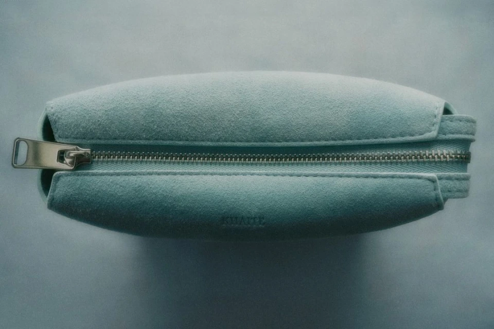 Close-up shot of a light blue suede cosmetic bag with silver zipper hardware, showcasing the fabric's texture and the design's clean lines.