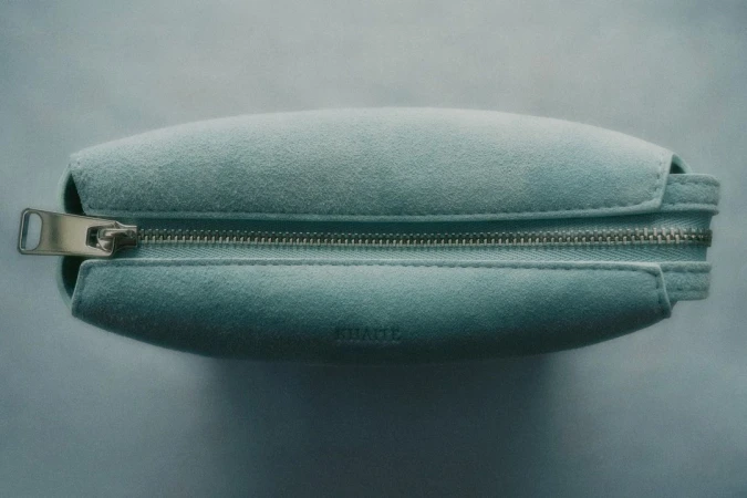 Close-up shot of a light blue suede cosmetic bag with silver zipper hardware, showcasing the fabric's texture and the design's clean lines.