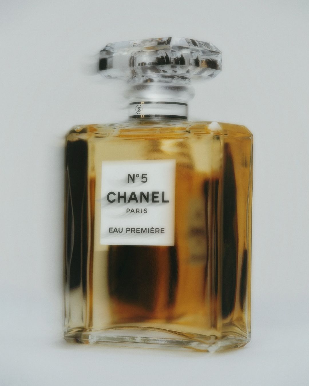 An amber-tinted Chanel N°5 Eau Première perfume bottle is centered on a light background, featuring a clear faceted stopper.