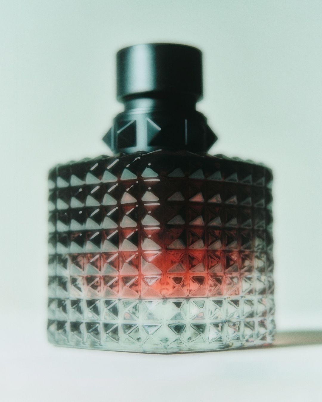 Studded glass perfume bottle with a black cap on a white surface against a pale green backdrop. Cosmetic product shot.