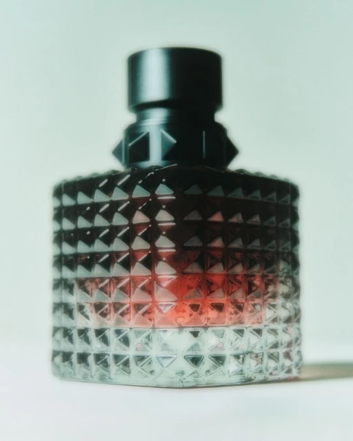 Studded glass perfume bottle with a black cap on a white surface against a pale green backdrop. Cosmetic product shot.