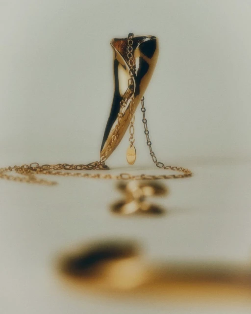 Close-up studio shot of a gold pendant and chain necklace on a cream surface. The gold jewelry is the main focus with a simple background.