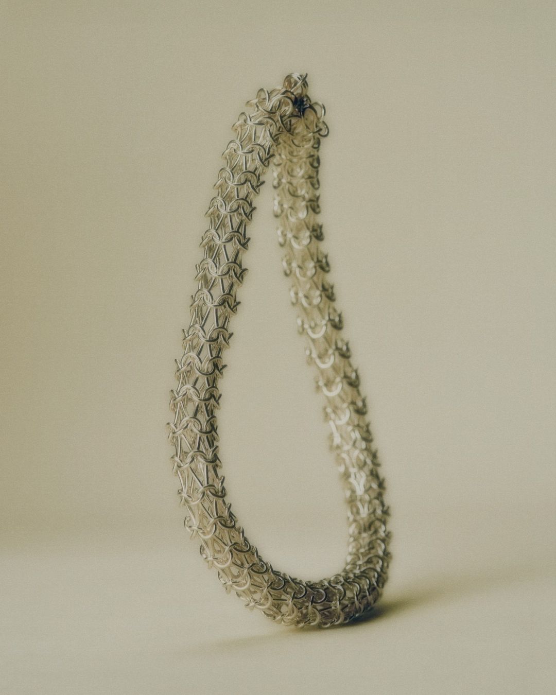 Close-up of a silver chainmail necklace, creating a teardrop shape against a soft, neutral background. The jewelry has an intricate pattern.