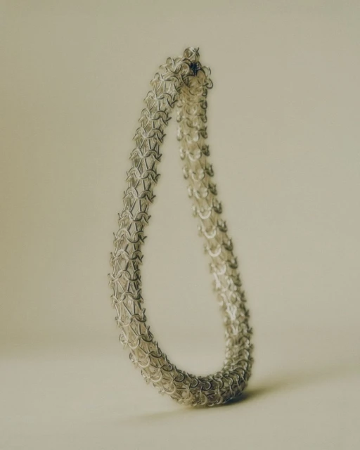 Close-up of a silver chainmail necklace, creating a teardrop shape against a soft, neutral background. The jewelry has an intricate pattern.