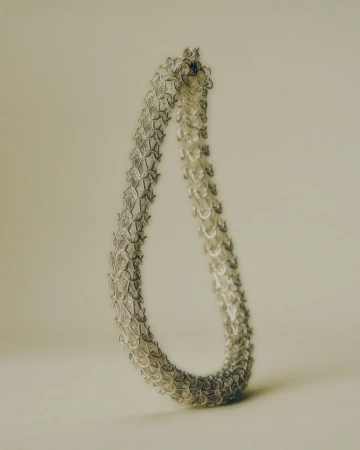 Close-up of a silver chainmail necklace, creating a teardrop shape against a soft, neutral background. The jewelry has an intricate pattern.