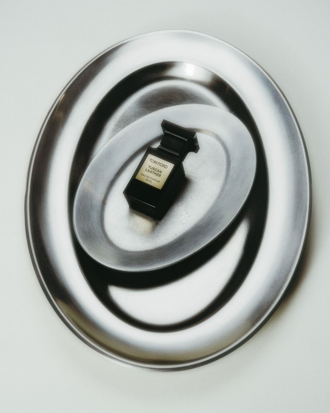 A black Tom Ford perfume bottle sits centered on a reflective silver plate, surrounded by a larger similar plate on a white surface.