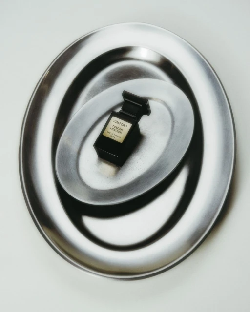 A black Tom Ford perfume bottle sits centered on a reflective silver plate, surrounded by a larger similar plate on a white surface.
