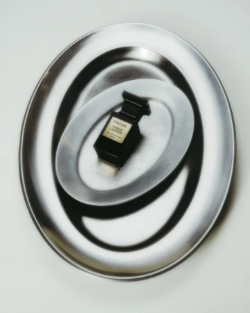 A black Tom Ford perfume bottle sits centered on a reflective silver plate, surrounded by a larger similar plate on a white surface.