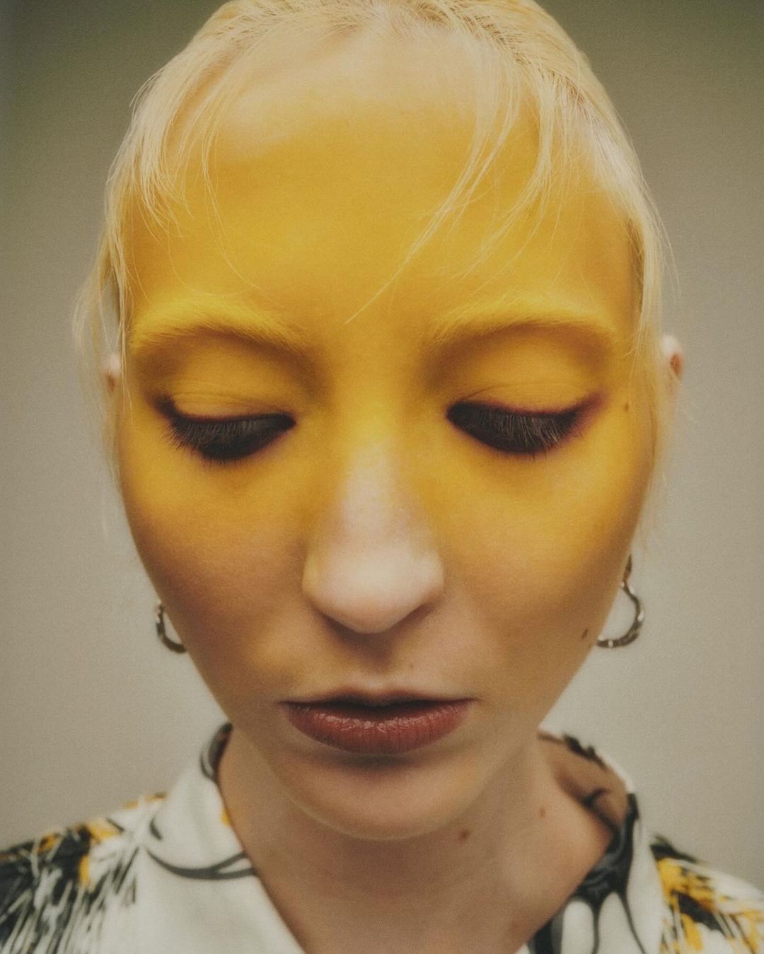 Portrait of a fair-skinned woman with blonde hair looking down, wearing makeup and patterned blouse with heart-shaped earrings.
