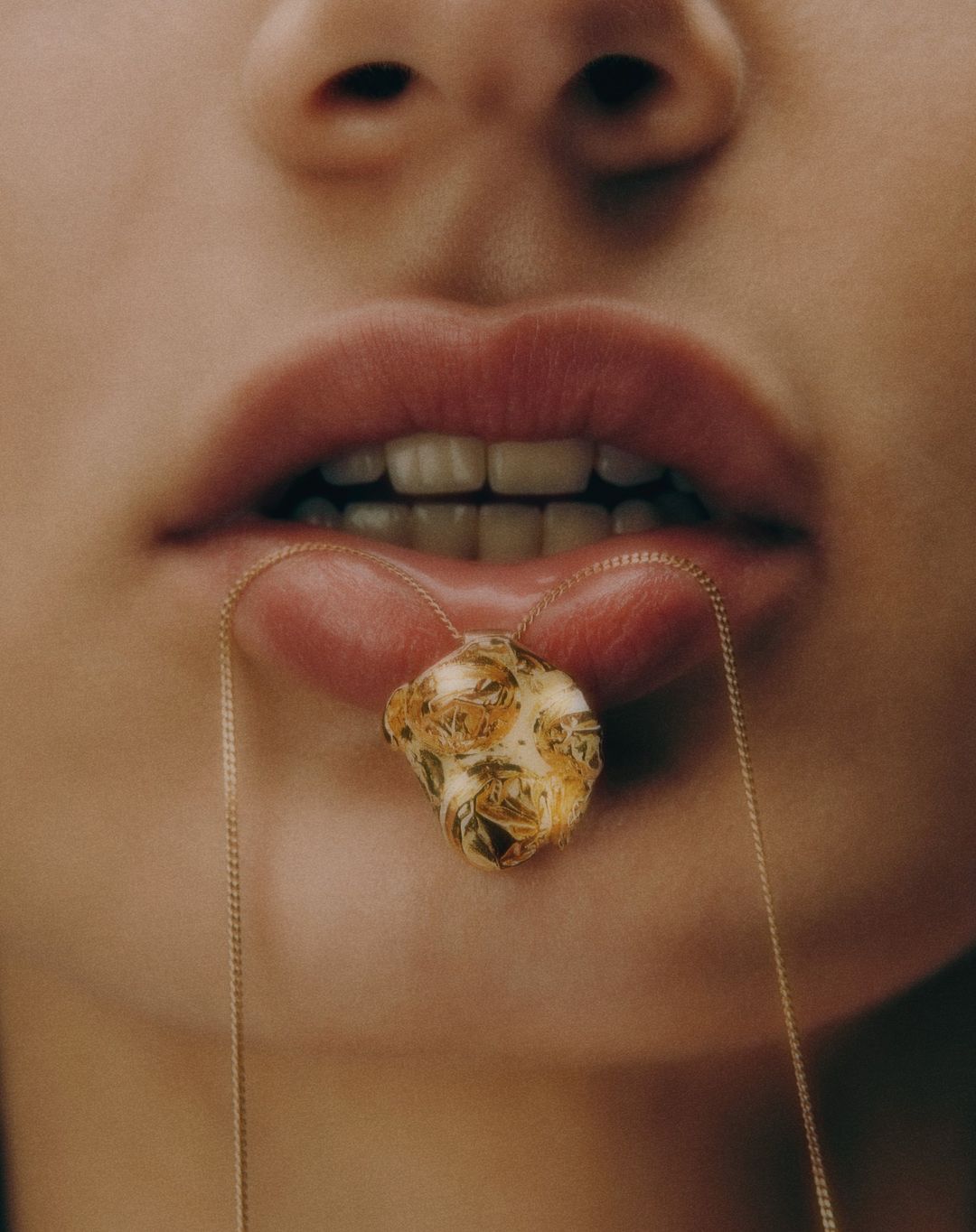 Close-up shot of a woman's mouth with parted lips, wearing a gold necklace with an ornate pendant in a studio setting. 