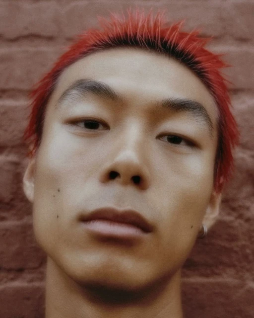 Close-up portrait of an Asian male with dyed red hair against a brick wall, showcasing a direct gaze and modern style in natural light.