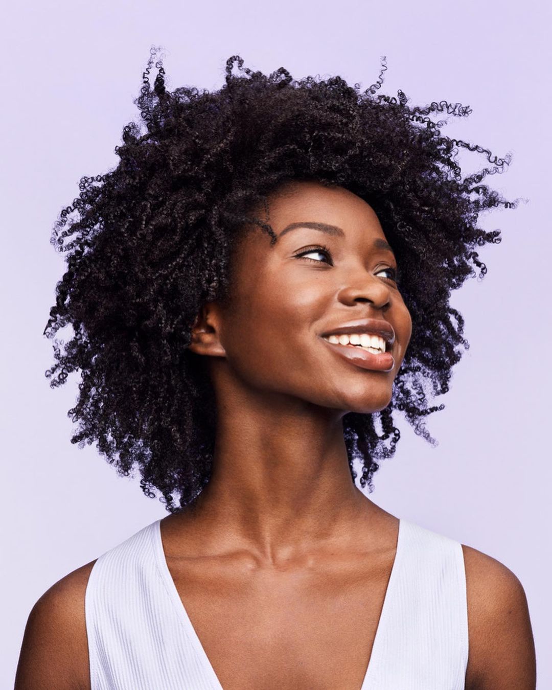 Smiling African American woman with voluminous curly hair poses in a sleeveless top against a light purple backdrop in a portrait.