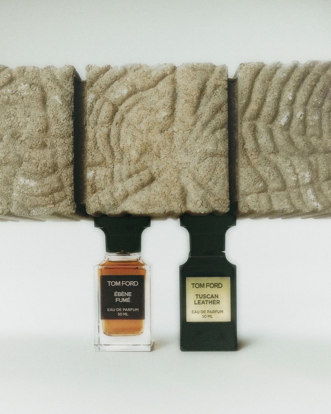 Two Tom Ford perfume bottles, one amber and one black, sit on a white surface beneath geometric stone blocks in a studio setting.