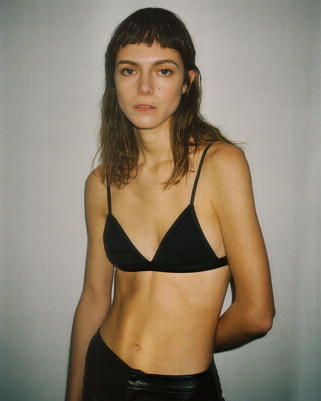 A slender woman with a fringe haircut poses in a black triangle bra and low-rise leather pants against a neutral white backdrop.
