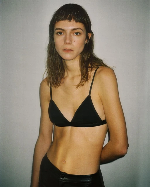 A slender woman with a fringe haircut poses in a black triangle bra and low-rise leather pants against a neutral white backdrop.