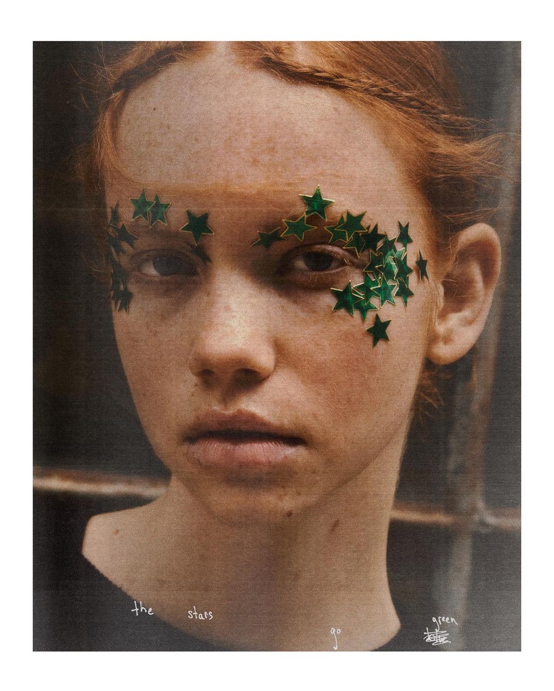 Close-up portrait of a young woman with freckles and red hair, adorned with green star-shaped face gems near her eyes.