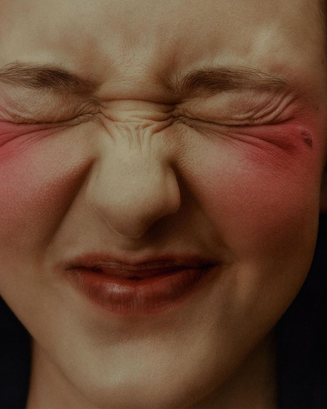 Close-up of an adult woman's face with eyes closed, red blush on cheeks, and a grimacing expression, set against a dark background.