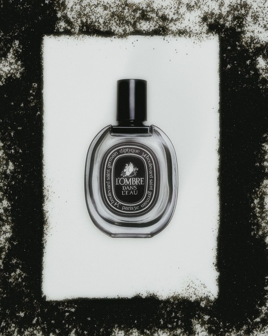 Studio shot of a Diptyque L'Ombre Dans L'Eau perfume bottle against a white backdrop with a black granular border.