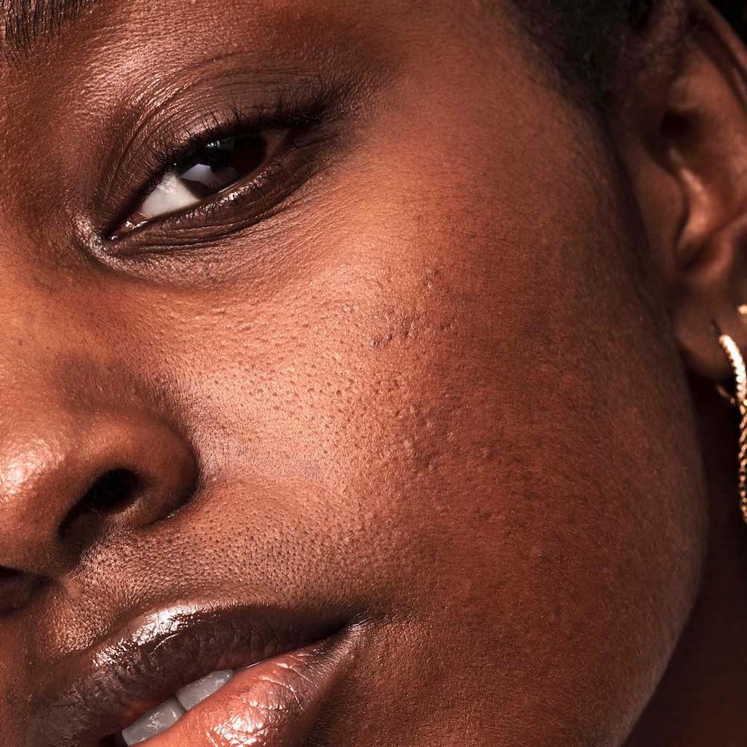 Close-up of an African American woman's face with smooth skin, showing makeup, an earring, and a subtle expression in a beauty shot.