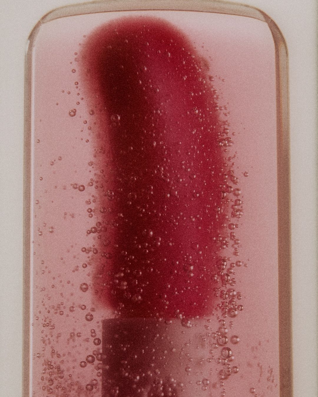 Close-up of lip gloss wand applicator inside tube, surrounded by light pink gloss liquid with small clear bubbles. Beauty product shot.