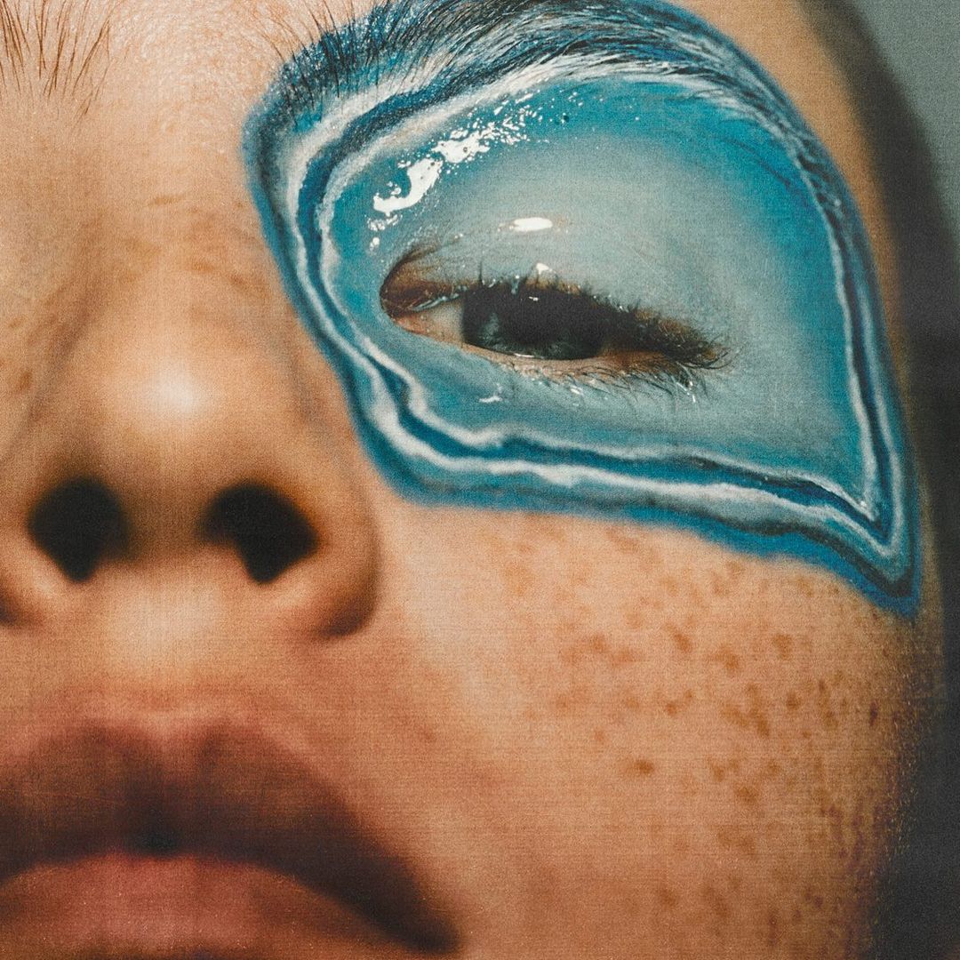 Close-up of a fair-skinned person with blue and white eye makeup extending from the eyelid to the eyebrow, highlighting artistic detail.