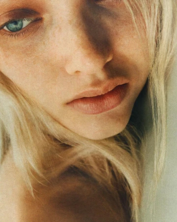 Close-up portrait of a fair-skinned woman with blonde hair and striking blue eyes, softly lit with focus on her natural features.