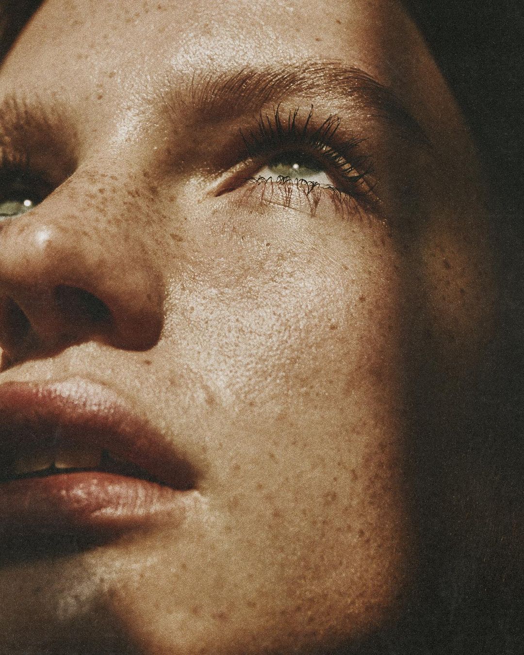 Close-up portrait of a young adult woman with freckles looking upward, focusing on her face, eyes, and skin details in natural lighting.