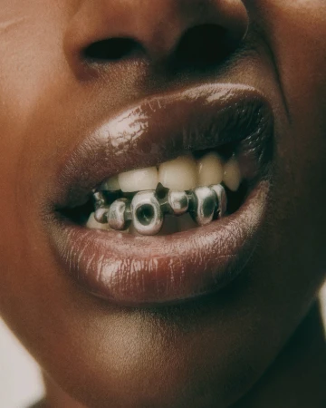 Close-up of a Black woman's mouth with silver tooth jewelry, showcasing glossy lips and dental accessories in a beauty or fashion context.