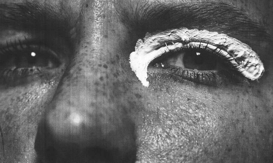 Close-up black and white shot of a person's face with white cream around their eye, highlighting eyelashes and eyebrow details.