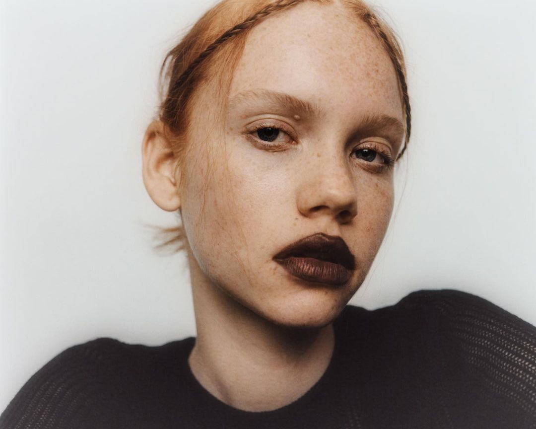 Close-up portrait of a fair-skinned young adult woman with reddish hair in a black top against a clean, pale background.