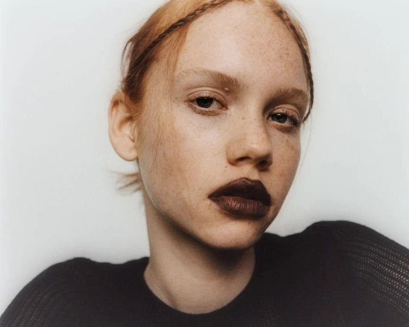 Close-up portrait of a fair-skinned young adult woman with reddish hair in a black top against a clean, pale background.
