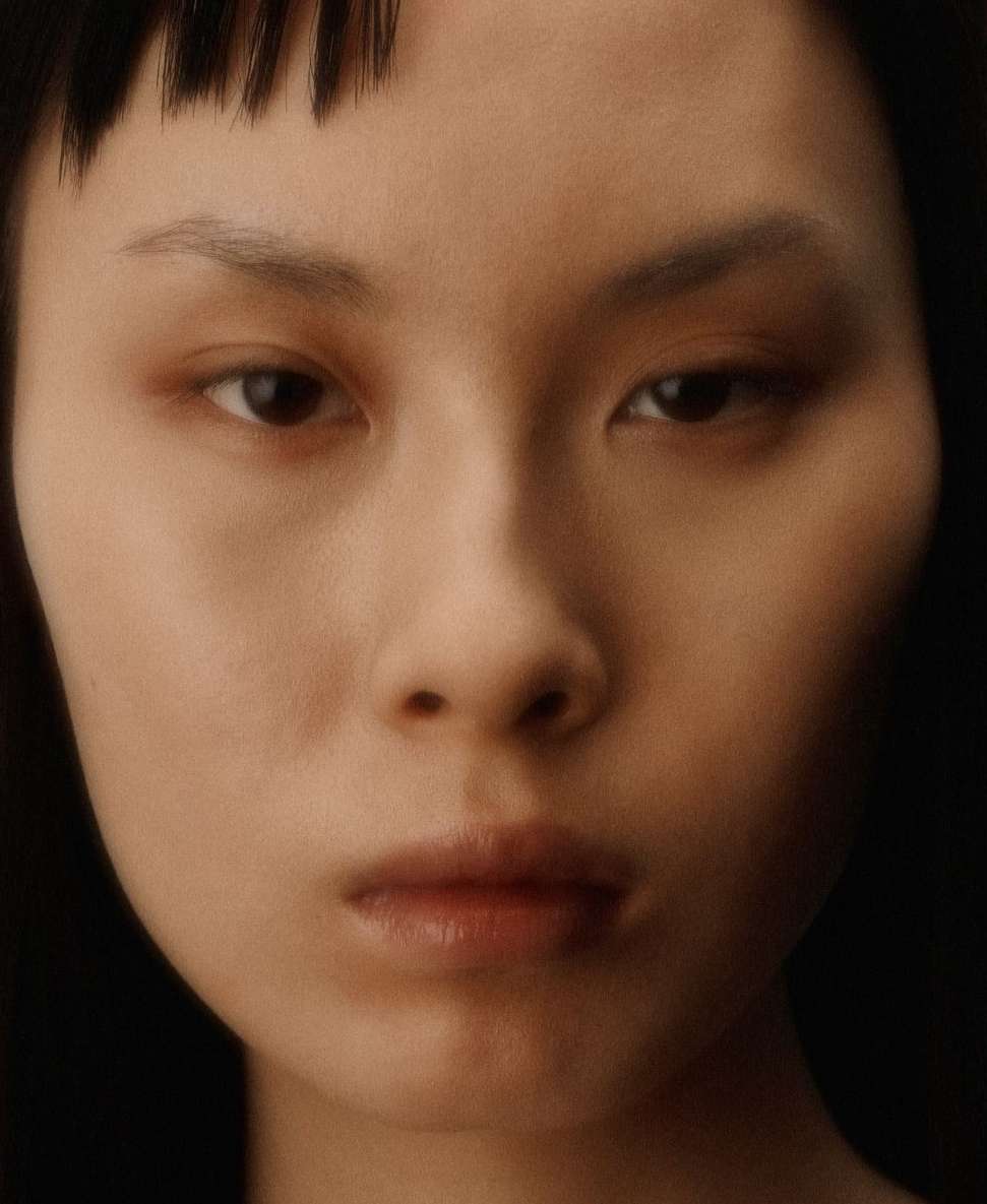 Close-up portrait of a young adult woman with dark hair and fair skin, softly lit in a photo studio against a dark backdrop.