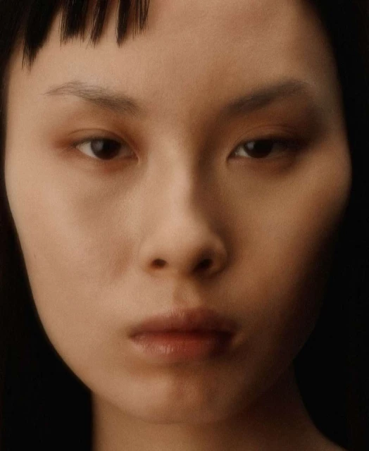 Close-up portrait of a young adult woman with dark hair and fair skin, softly lit in a photo studio against a dark backdrop.
