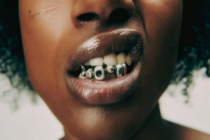 Close-up studio shot of a Black woman's mouth with a silver grill. The lips are glossy and the background is a neutral color.