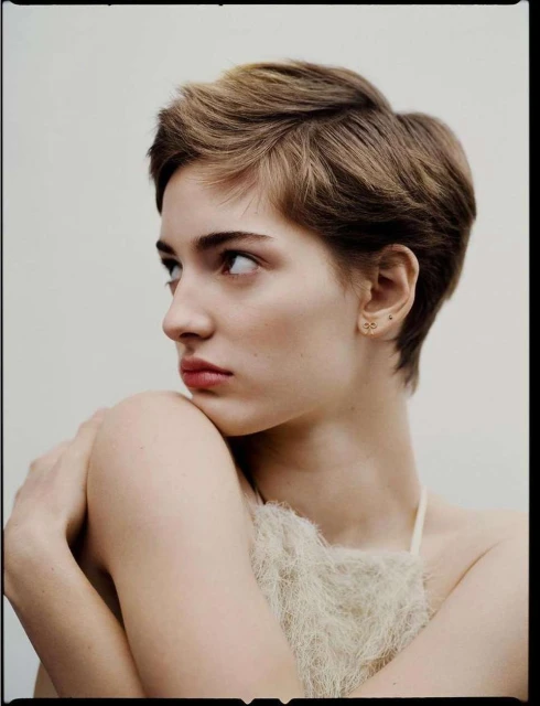 Close-up portrait of a young adult female with a short haircut, fair skin, bare shoulders, wearing small gold earrings, in a studio setting.