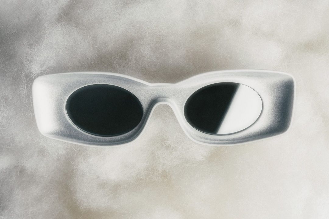 Elevated studio shot of futuristic rimless sunglasses with gray frames and dark lenses resting on a textured white fluffy surface.