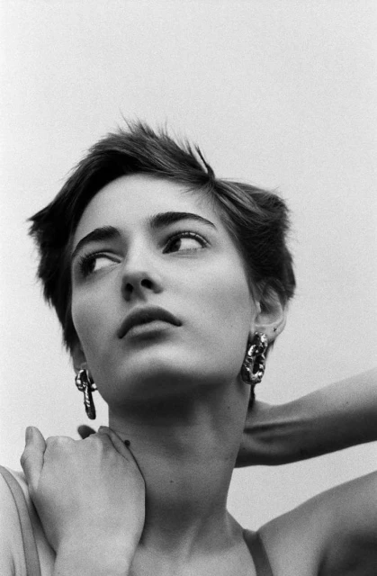 Portrait of a woman with short dark hair and statement earrings, looking up and to the side against a light background in black and white.