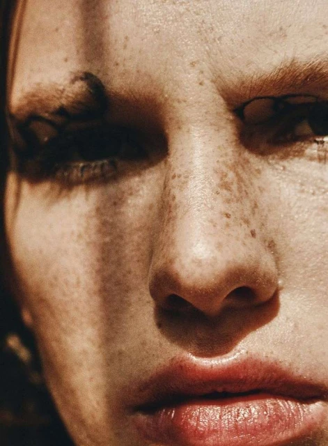 Close-up portrait of an adult female face with freckles and red lips, partially lit with shadows, in a beauty and fashion style.