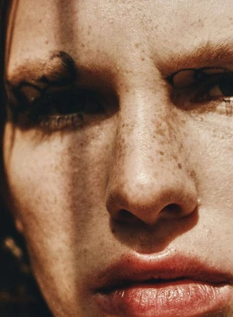Close-up portrait of an adult female face with freckles and red lips, partially lit with shadows, in a beauty and fashion style.