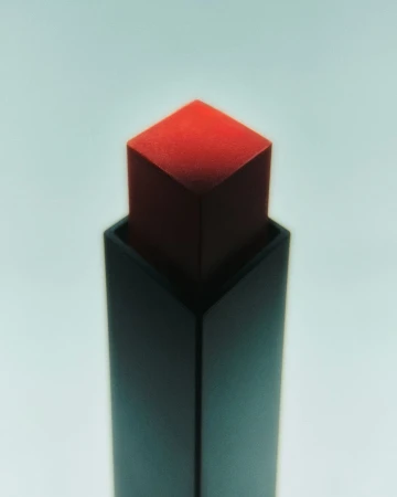 Close-up of a square red lipstick component extended from a dark matte square tube against a pale gradient background.