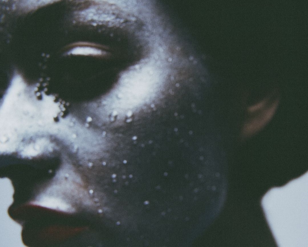 Close-up portrait of a person with silver body paint and water droplets, focusing on the face and side profile.